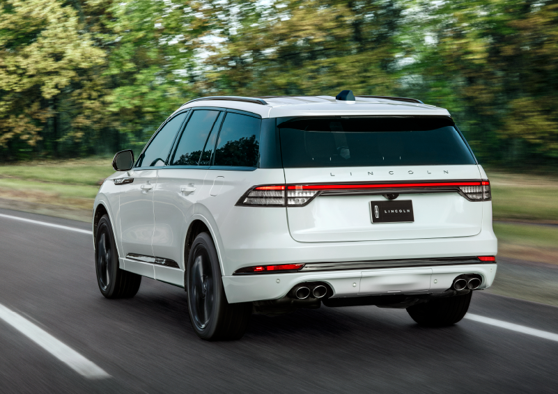 A white 2025 Lincoln Aviator® SUV is being driven away down a road lined with colorful trees. | Planet Lincoln Love Field in Dallas TX