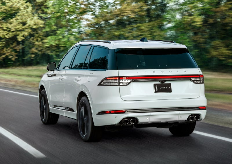 A white 2026 Lincoln Aviator® SUV is being driven away down a road lined with colorful trees. | Planet Lincoln Love Field in Dallas TX